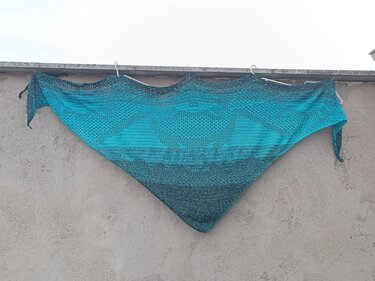 Gehäkeltes Dreieckstuch in Türkis und Grau mit zentralem Herz-Lochmuster, an Wand aufgehängt