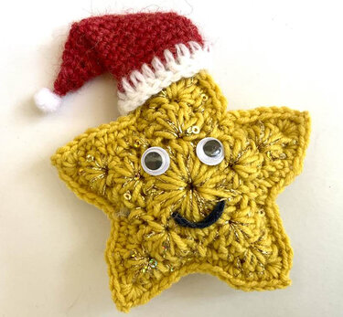 Gehäkelter gelber Stern mit Wackelaugen, gesticktem Mund und roter Weihnachtsmütze