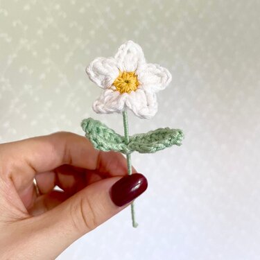 Hand hält gehäkelte weiße Blume mit gelber Mitte und grünen Blättern