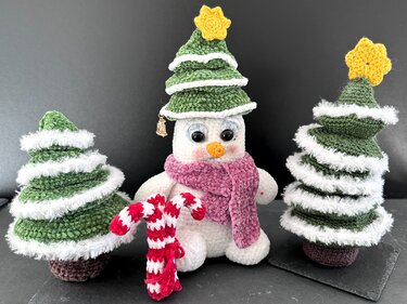Gehäkelter Schneemann mit grüner Tannenmütze, rosa Schal und gehäkelter Zuckerstange, flankiert von zwei kleinen gehäkelten Weihnachtsbäumen mit weißen Borten.