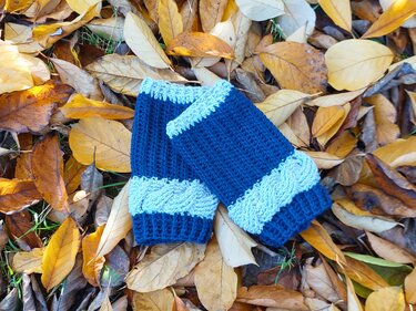 Zwei blaue gehäkelte Armstulpen mit Zopfmuster auf gelb-braunem Herbstlaub.