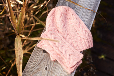 Rosa gestrickte Mütze und passender Loop mit Zopfmuster auf einer Holzbank