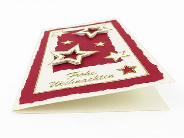 Nahaufnahme einer DIY-Weihnachtskarte mit reliefartigen 3D-Sternen, rotem Einleger und goldenen Glitzerrändern