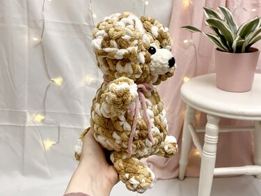 Hand hält einen flauschigen, gehäkelten Amigurumi-Hund in Braun und Weiß mit rosa Schleife