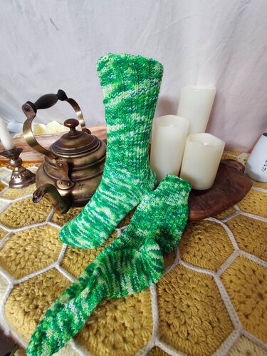 Gestrickte grüne Socken (meliert) auf gelbem Häkeldeckchen neben Messingkanne und Kerzen