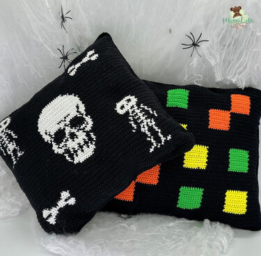 Zwei gehäkelte schwarze Kissen: eines mit Totenkopf- und Skelettmustern, das andere mit grünen, orangen und gelben Quadraten, auf Spinnennetz-Deko