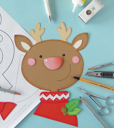 Ausgeschnittener Rentierkopf aus Papier mit rotem Pullover, daneben Schnittmuster und Bastelwerkzeuge auf blauem Tisch.