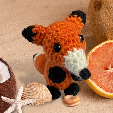kleiner gehäkelter oranger Fuchs-Amigurumi mit weißer Schnauze und schwarzen Pfoten neben Muscheln und einer Grapefruit‑hälfte