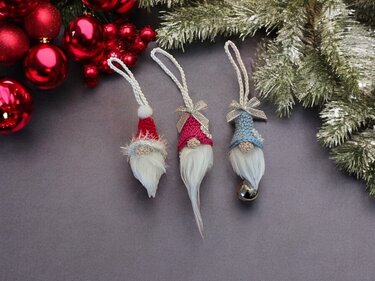 Nahaufnahme von drei gehäkelten Wichteln mit farbigen Mützen, weißen Bärten, Aufhängeschlaufen und einer kleinen Glocke; umgeben von Weihnachtsdekoration.