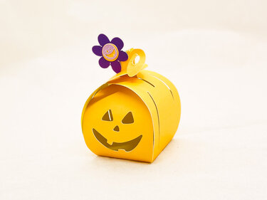 Gelbe Papier-Geschenkbox in Kürbisform mit ausgeschnittenem Jack‑o‑Lantern-Gesicht und lila Blütenverschluss