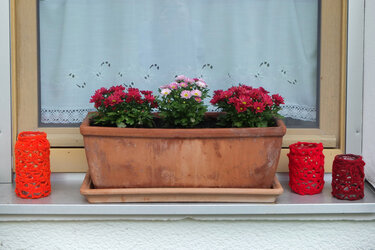 Terracotta-Blumenkasten mit roten und rosa Chrysanthemen und gehäkelten Windlichtern auf einer Fensterbank vor einer weißen Gardine.
