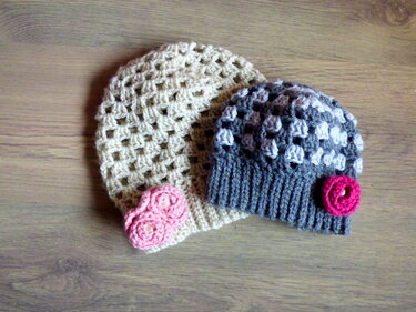 Zwei gehäkelte Mützen auf Holz: eine cremefarbene mit rosa Rosen, eine graue mit pinker Rose.