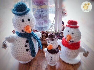 Drei gehäkelte Amigurumi-Schneemänner mit blauer, brauner und roter Mütze und Schal auf Holzoberfläche.