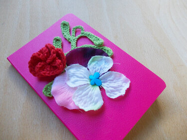 Pinkes Notizbuch mit roter Häkelrose, weiß-rosafarbener Stoffblüte und grünen Häkelblättern