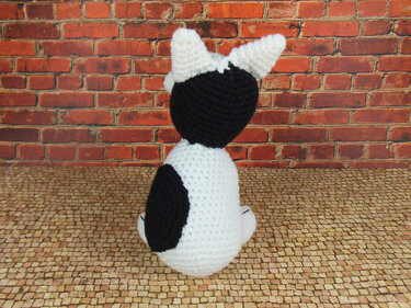Schwarz-weißes Amigurumi-Kätzchen, Rückansicht vor roter Backsteinwand
