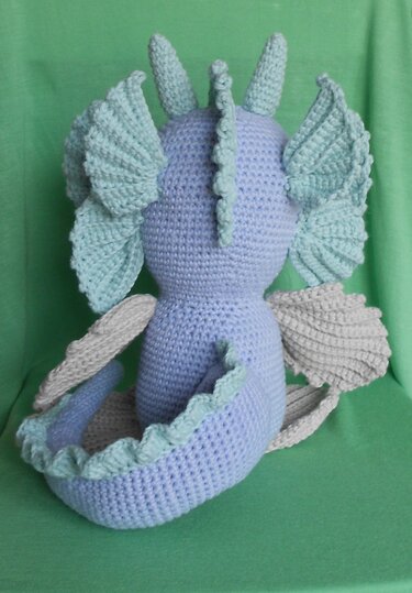 Rückansicht eines gehäkelten Amigurumi-Drachens in Hellblau mit mintgrünen Details und hellgrauen Flügeln vor grünem Hintergrund.