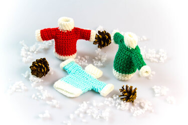 Drei gehäkelte Mini-Pullover (rot, grün, hellblau) mit weißen Bündchen, Tannenzapfen und Kunstschnee