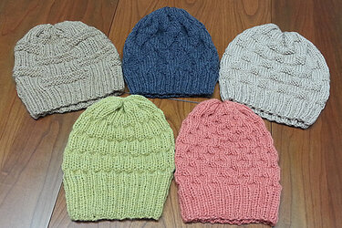 Fünf gestrickte Beanies in Sand, Dunkelblau, Hellgrau, Lindgrün und Koralle auf Holzuntergrund, mit Rippenbündchen und strukturiertem Strickmuster