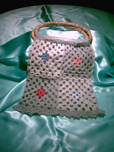 Gehäkelte graue Henkeltasche mit Lochmuster, kleinen roten und blauen Blüten und runden Bambusgriffen auf türkisfarbenem Stoff.