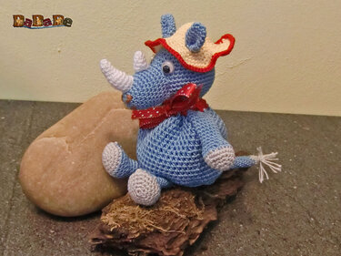 Blaues gehäkeltes Nashorn-Amigurumi mit weißem Horn, cremefarbenem Hut und roter Schleife, sitzend auf Naturmaterialien