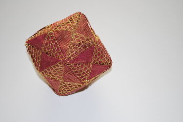 Detailaufnahme eines geometrischen Faltobjekts aus bordeauxrotem Stoff und goldenem Netzgewebe.