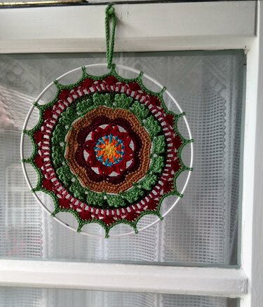 Gehäkeltes rundes Mandala in Rot-, Grün- und Brauntönen, eingespannt in weißen Metallring, aufgehängt vor einem Fenster mit weißer Gardine