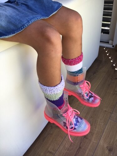 Beine in Jeansrock mit bunten gehäkelten Socken in transparenten Stiefeln mit pinken Schnürsenkeln.