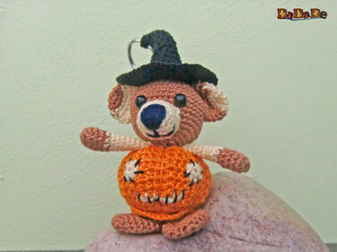 Gehäkelter kleiner Bär mit braunem Fell, blauem Näschen, schwarzem Hexenhut und orangefarbenem Kürbiskörper mit Jack‑O'‑Lantern‑Gesicht, an einem Schlüsselring, sitzend auf einem Stein.