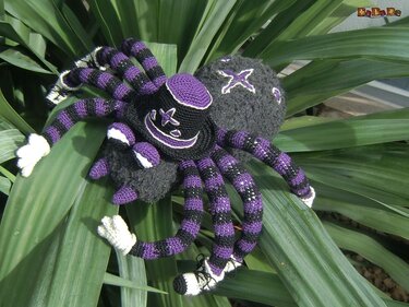 Gehäkelte Amigurumi-Spinne in Lila und Schwarz mit gestreiften Beinen auf grünen Blättern