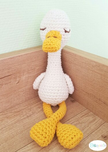 Gehäkelte weiße Ente mit gelbem Schnabel, verschränkten gelben Füßen und gestickten geschlossenen Augen, sitzt in einer Holzecke
