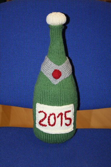 Gestrickte grüne Sektflasche mit weißem Flaschenkorken, grauem Kragen und Etikett '2015'.
