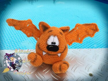 Oranges gehäkeltes Amigurumi-Fledermausmonster auf metallischem Untergrund vor blauem Hintergrund
