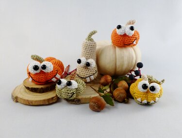Gehäkelte Amigurumi-Kürbismonster mit großen Augen und Zähnen auf Holzscheiben, umgeben von Nüssen und einem weißen Kürbis.
