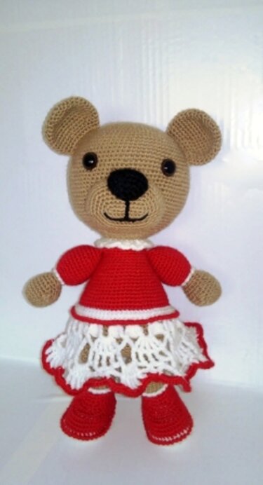 Gehäkelter beiger Amigurumi-Teddybär mit rotem Oberteil, weißem Spitzenrock und roten Schuhen, vor einem hellen, neutralen Hintergrund.