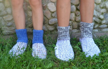 Zwei Paare gestrickter Hausschuhe mit hohem Rippbündchen in Blau und Grau auf Gras vor einer Steinmauer