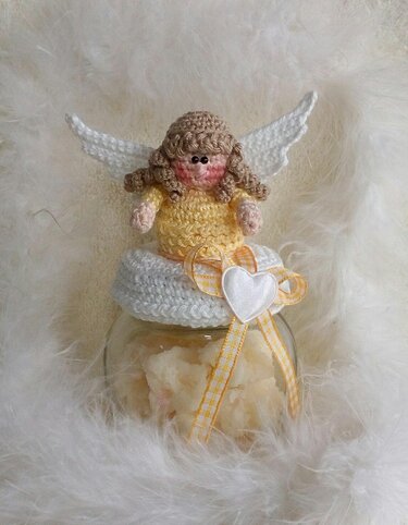 Gehäkelter Engel in gelbem Kleid mit weißen Flügeln auf dekoriertem Glas vor weißem Federhintergrund