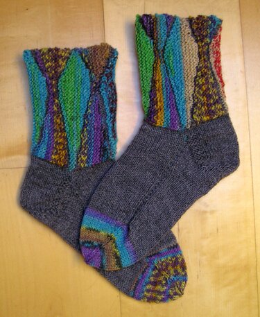 Ein Paar handgestrickte Damensocken mit grauem Fußteil und bunten Manschetten, liegend auf hellem Holzuntergrund.