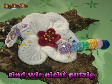 Gehäkelte weiße Blume mit rotem Zentrum, kleiner roter Blüte, weiß-violetter Schmetterling und mehrfarbige Raupe auf Baumrinde mit Moos.
