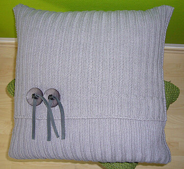 Hellgraues Strickkissen mit vertikalem Rippenmuster und zwei Holzknöpfen mit grauen Bändern
