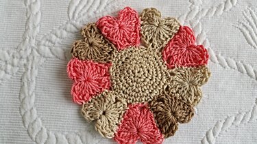 Runder gehäkelter Untersetzer mit Herzmotiven in Rosa, Beige und Braun auf weißer Stoffunterlage