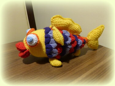 Gehäkelter Amigurumi-Fisch in Gelb mit mehrfarbigen Schuppen, Seitenansicht auf Holzfläche