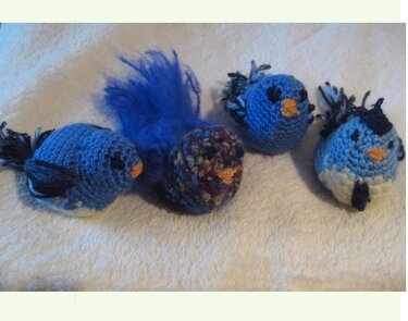 Vier gehäkelte Amigurumi-Vögel in Blautönen, einer mit bunt gemustertem Rumpf und flauschigem blauem Schwanz auf hellem Tuch