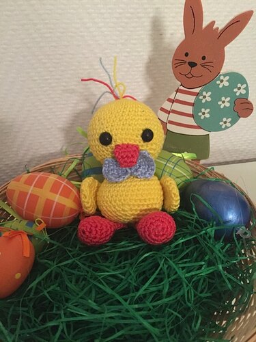Gelbes gehäkeltes Amigurumi-Küken mit blauer Fliege sitzt im Korbnest auf grünem Ostergras zwischen bunten Ostereiern
