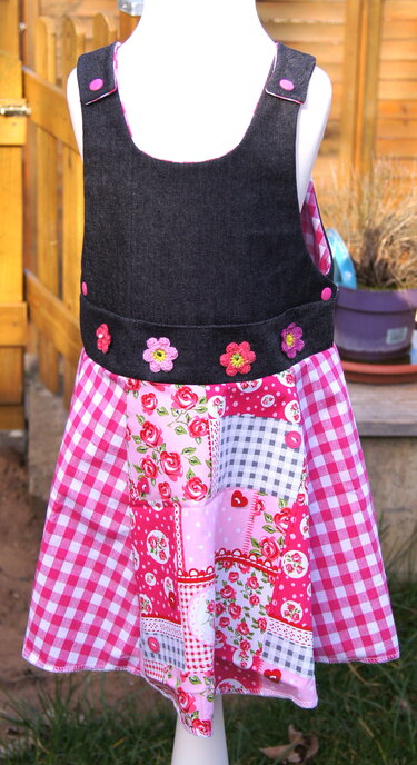 Kinder-Latzkleid mit dunklem Oberteil, gehäkelten Blumen am Bund und rosa-weißem Patchwork-Rock auf einer Schaufensterpuppe