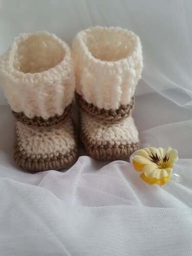 Zwei gehäkelte Babyschuhe in Creme und Beige mit umgeschlagenem Bündchen auf weißem Tüll, daneben eine gelbe Stoffblume