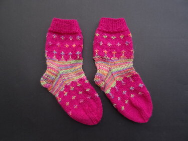 Ein Paar fuchsiafarbene gestrickte Strümpfe mit bunten Streifen und kleinen Mustern auf dunklem Untergrund.