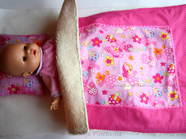 Baby doll tucked into a pink mushroom-and-flower blanket with polka-dot trim and cream plush underside and a matching pillow.