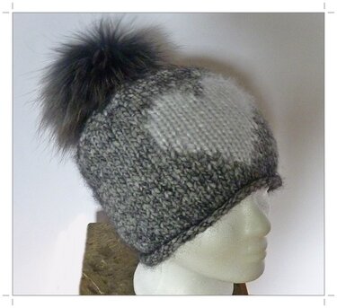 Grau-weiß melierte Strickmütze mit Strukturmuster und großem dunklem Bommel auf Schaufensterpuppe.
