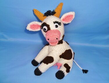 Gehäkelte Amigurumi-Kuh mit braunen Flecken, rosa Schnauze und gelben Hörnern vor blauem Hintergrund