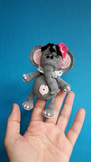 Gehäkelte graue Elefanten-Fingerpuppe mit rosa Details und Herz auf einer Hand vor blauem Hintergrund